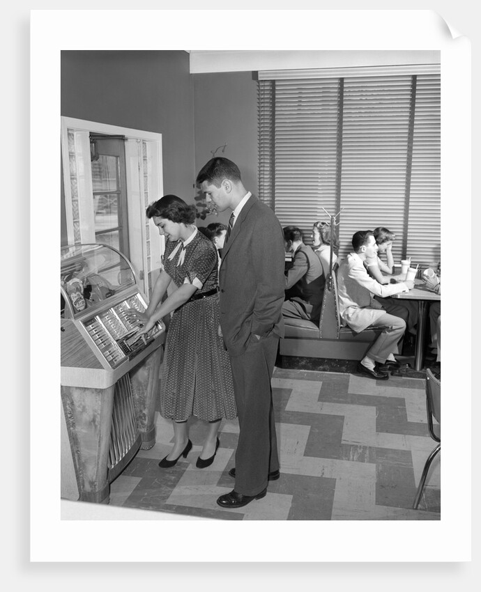 1950s teen couple playing juke box in malt shop with other teens in booths by Anonymous
