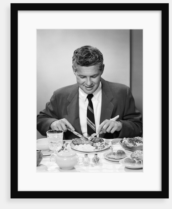 1950s smiling man in suit and tie sitting at table holding knife and fork eating dinner by Anonymous
