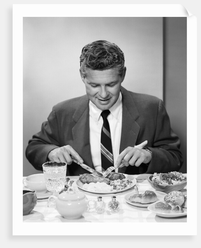 1950s smiling man in suit and tie sitting at table holding knife and fork eating dinner by Anonymous