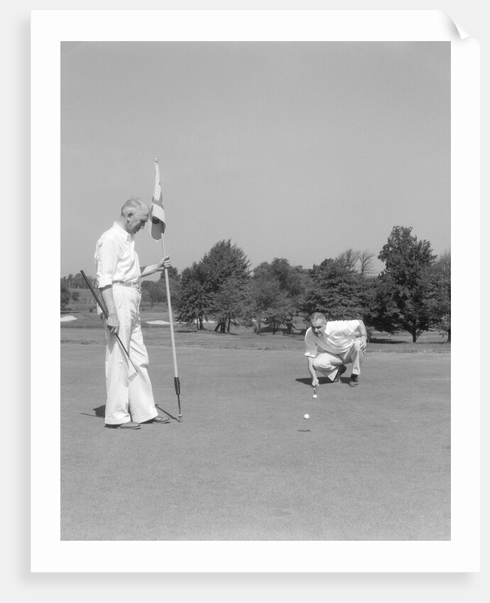 1930s 1940s elderly men on golf green one holding flag the other kneels lining up his putt by Anonymous