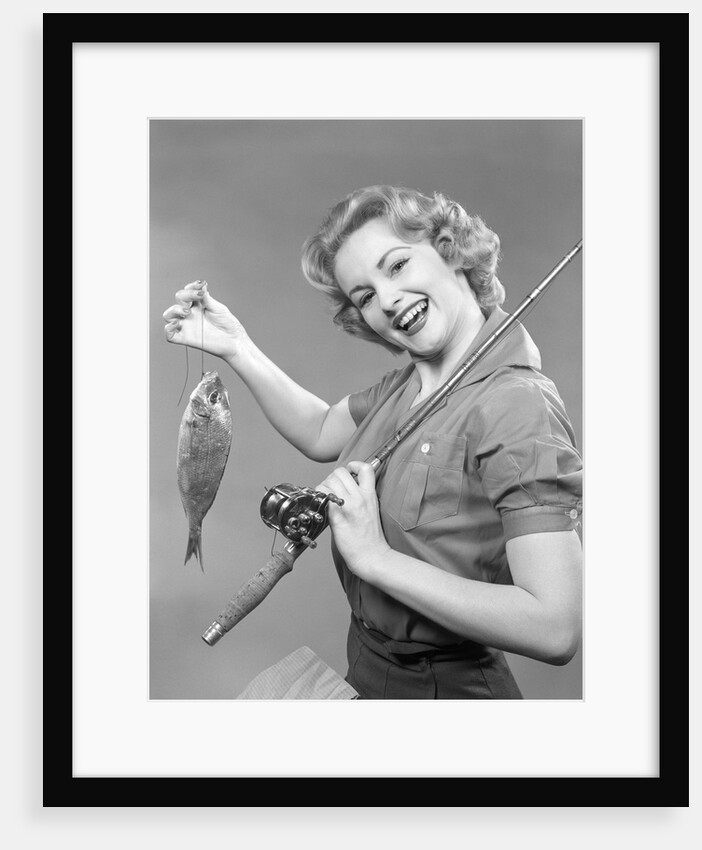 1950s smiling woman with a fishing rod over her shoulder holding up a fish looking at camera by Anonymous