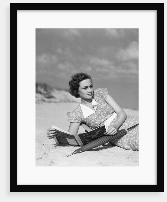 1930s woman leaning in wooden chair on beach wearing printed dress holding magazine looking thoughtful by Anonymous