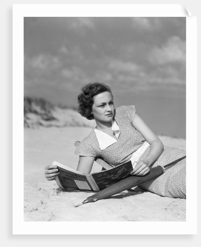 1930s woman leaning in wooden chair on beach wearing printed dress holding magazine looking thoughtful by Anonymous