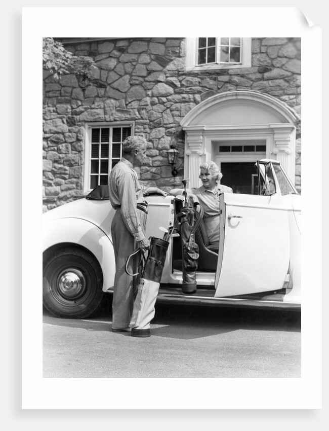 1940s senior retired couple loading golf clubs into white convertible car in front stone house by Anonymous