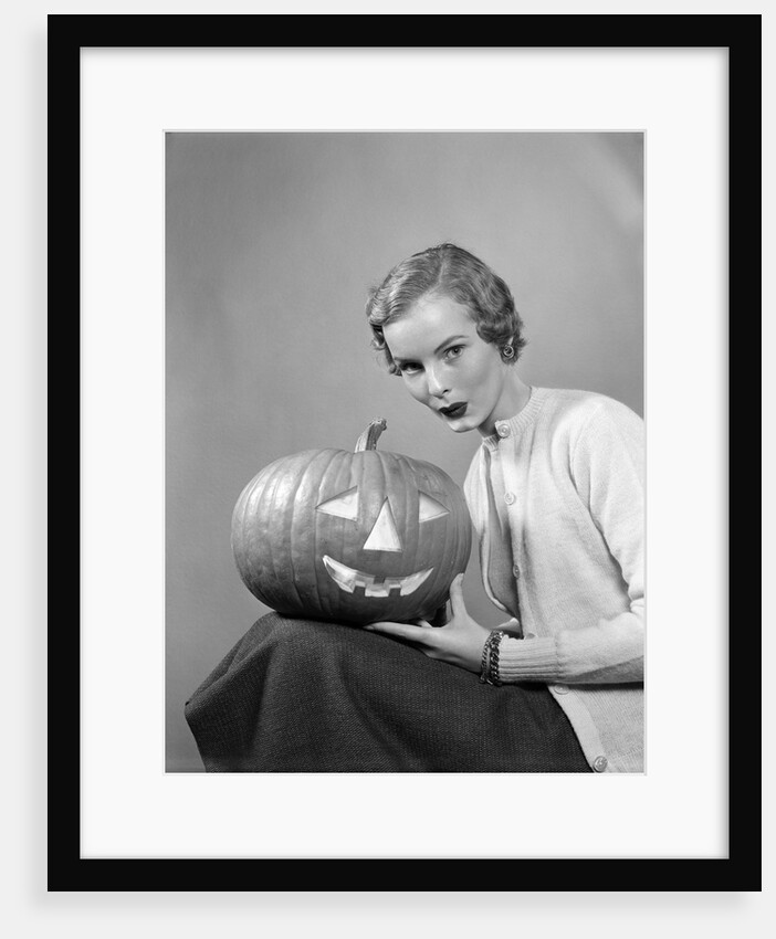 1950s woman wearing sweater holding carved pumpkin jack-o-lantern on her lap by Anonymous