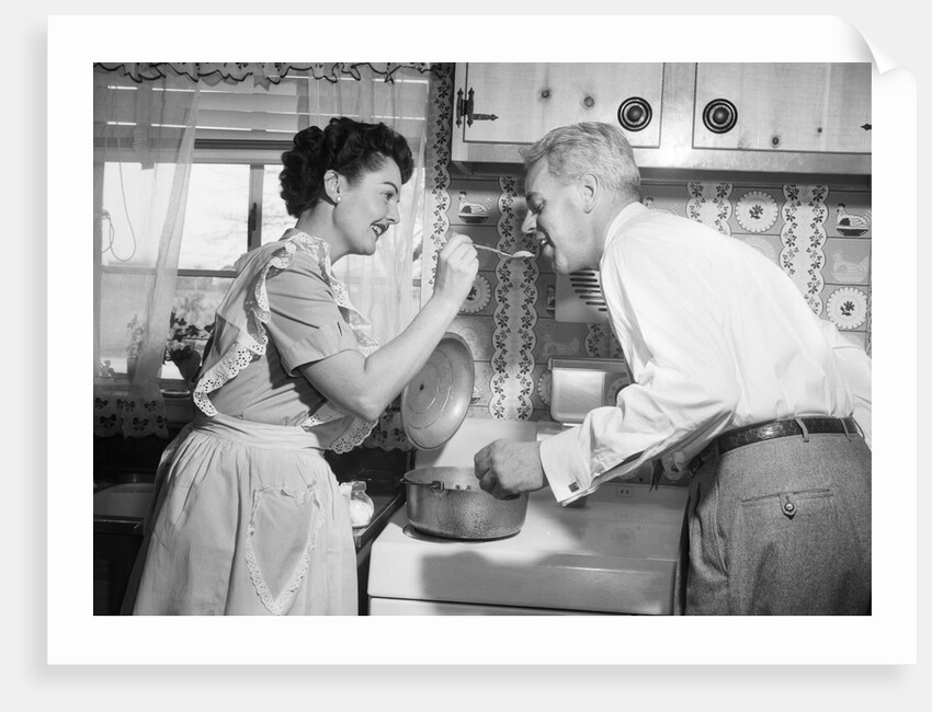 1950s housewife in kitchen having husband taste food on stove by Anonymous