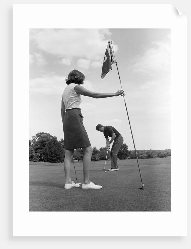 1970s couple playing golf man putting woman holding flag by Anonymous