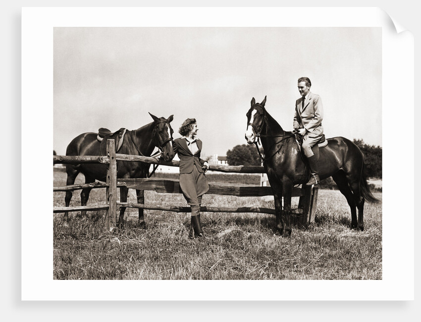 1930s 1940s couple in riding gear man riding horse woman standing by wooden fence by Anonymous