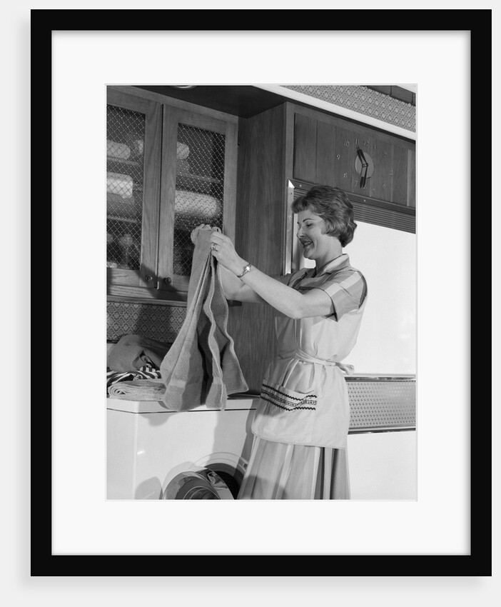 1960s woman folding laundry towels on dryer in kitchen by Anonymous
