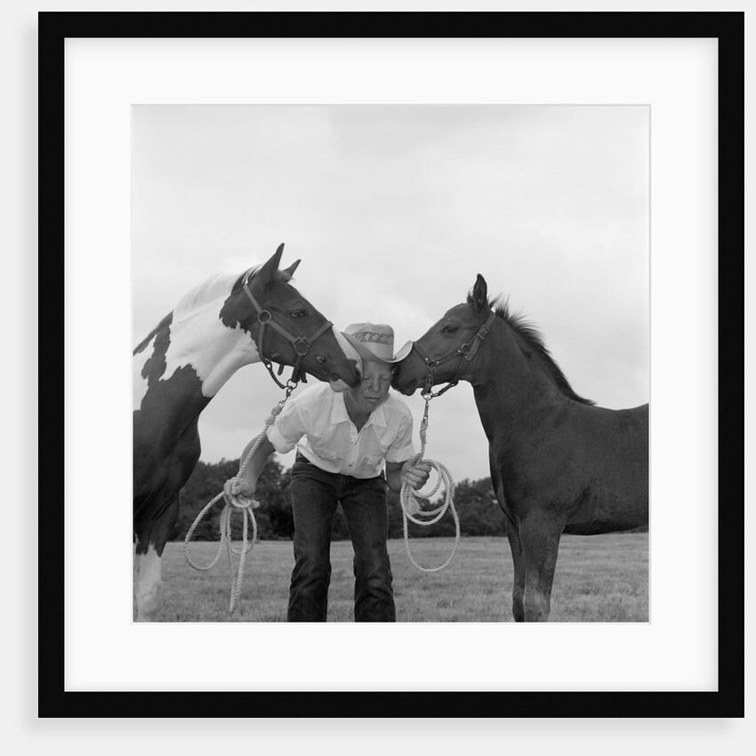 1960s boy wearing cowboy hat holding ropes reins harness halter of 2 horses one on either side of his face kissing him funny by Anonymous