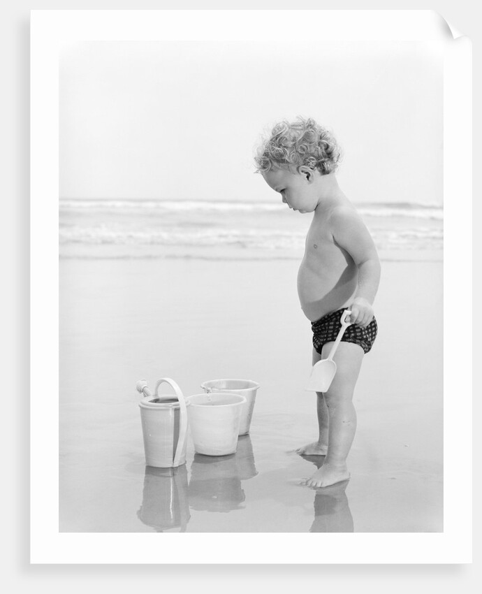 1960s 1970s child toddler standing in sand water bucket shovel by Anonymous