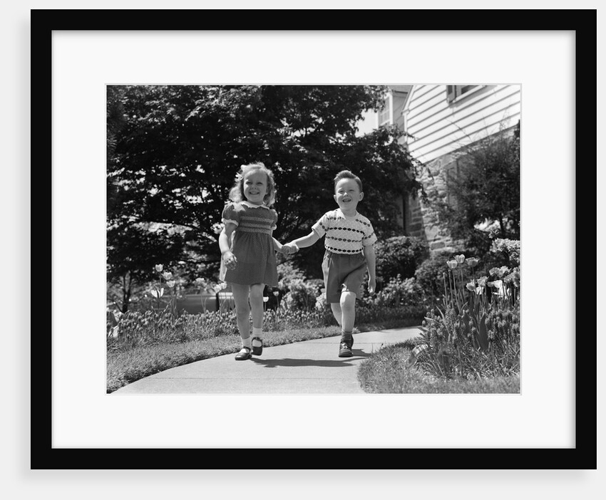 1950s two children holding hands smiling walking along sidewalk by Anonymous