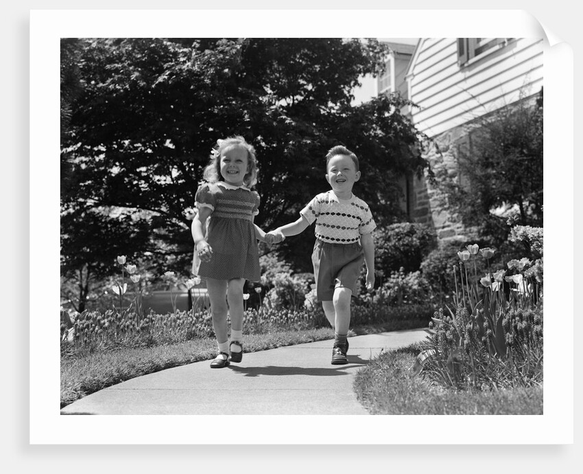 1950s two children holding hands smiling walking along sidewalk by Anonymous