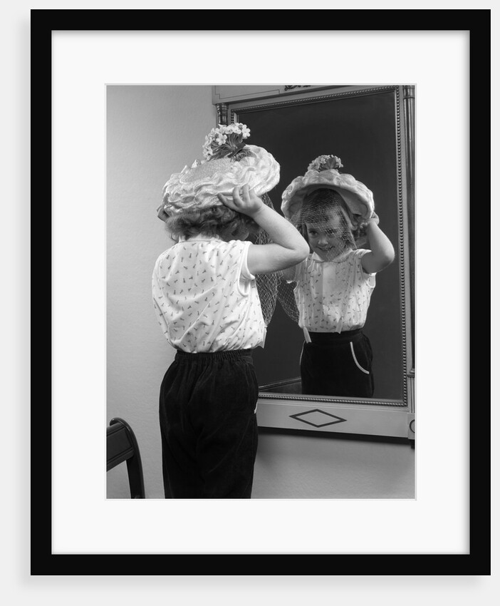 1950s little girl trying on hat looking into mirror reflection by Anonymous