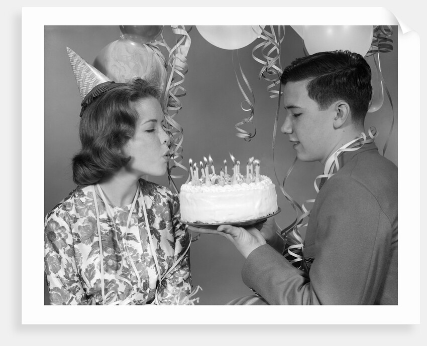 1960s teenage girl blowing out candles on birthday cake held by boy by Anonymous