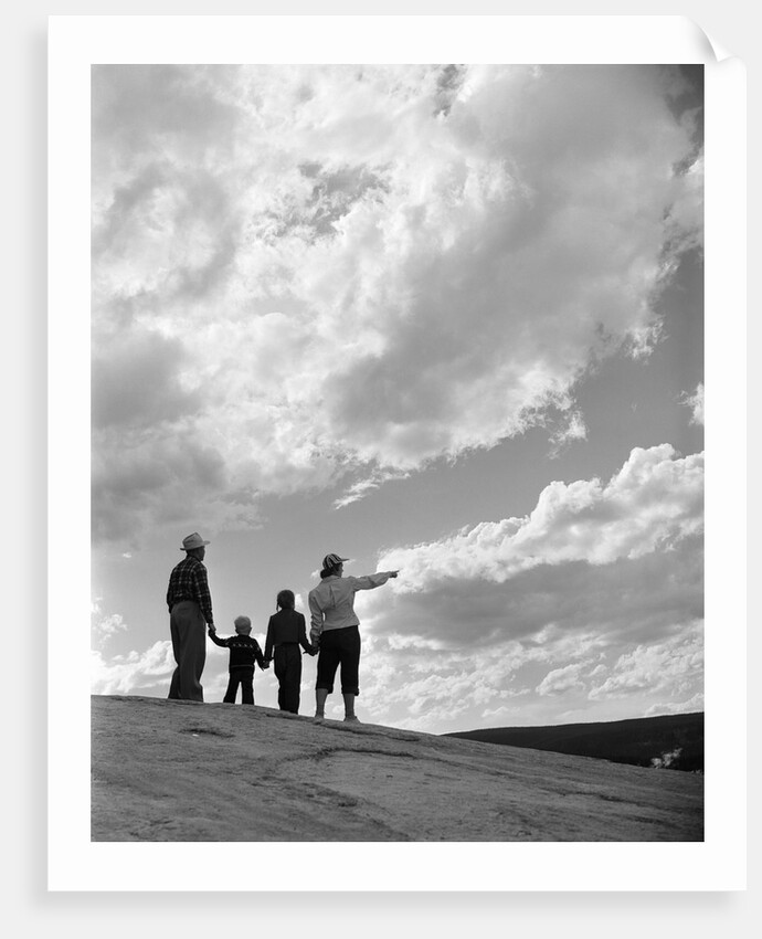 1950s 1960s family of four silhouetted on top rocky hill pointing at clouds in sky by Anonymous