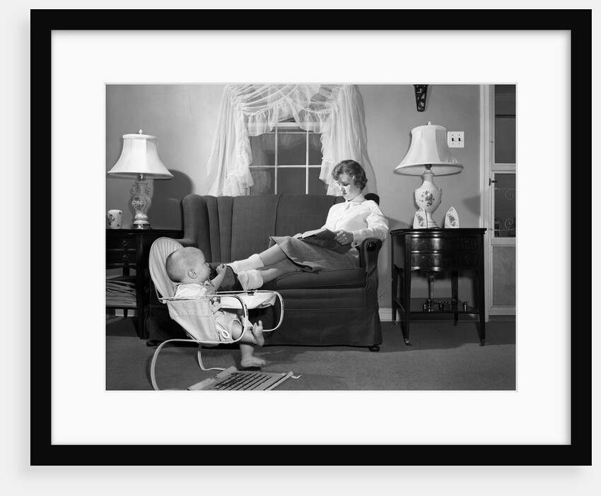 1950s teen babysitter seated on sofa reading schoolbook while baby in bouncy sling chair grabs her propped up foot by Anonymous