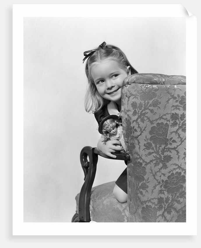 1940s little girl holding doll peeking around side of chair by Anonymous