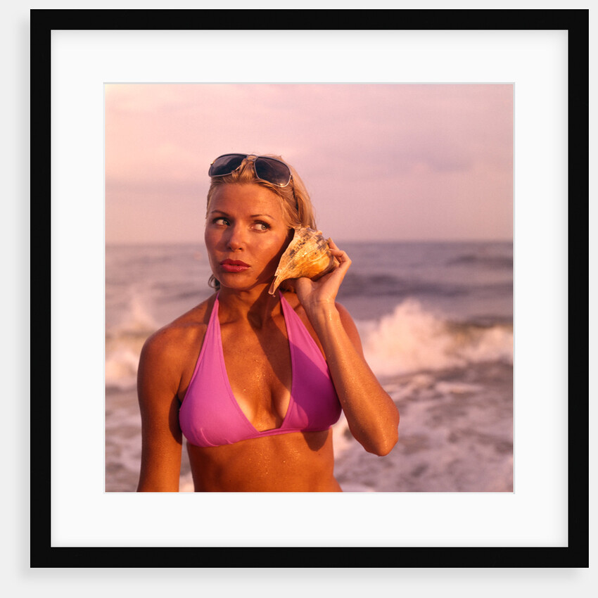 1970s pouting sexy woman in lavender bikini sunglasses near beach surf holding seashell to ear listening to ocean by Anonymous
