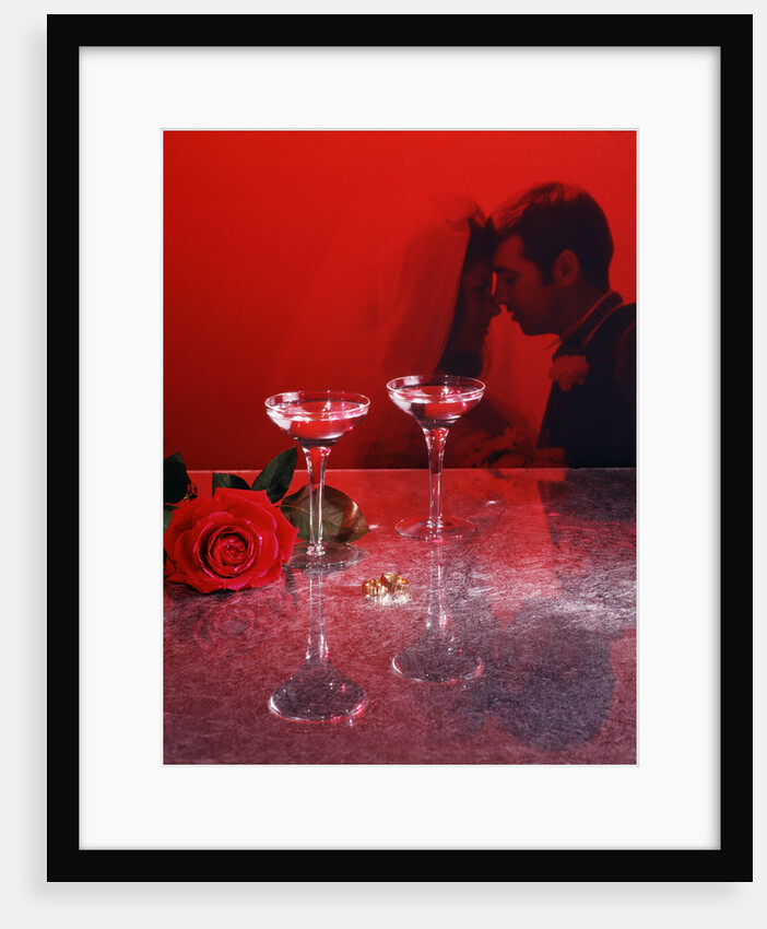 1960s champagne flutes with rose and wedding rings in red filter with couple man woman bride groom in background by Anonymous