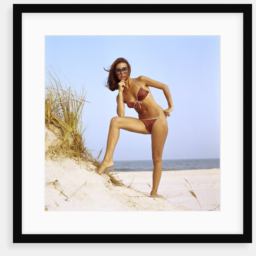 1970s woman wearing two piece string bikini bathing suit sunglasses posing by grassy sand dune at beach by Anonymous
