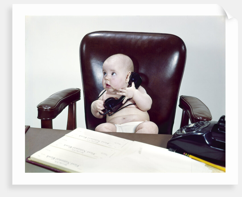 1960s chubby baby sitting in leather chair at office desk holding telephone by Anonymous