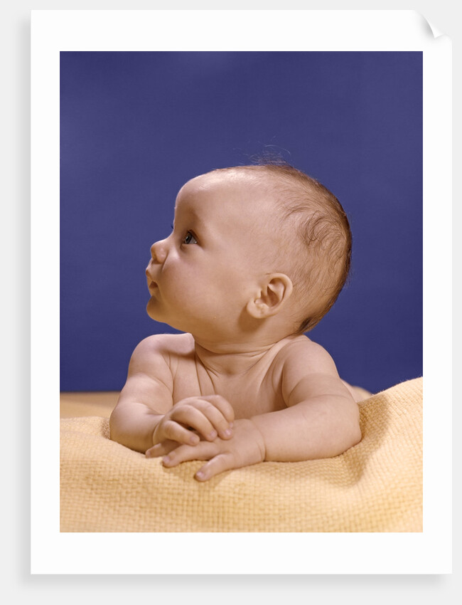 1960s portrait of baby on yellow blanket resting on folded arms head turned to right looking up by Anonymous