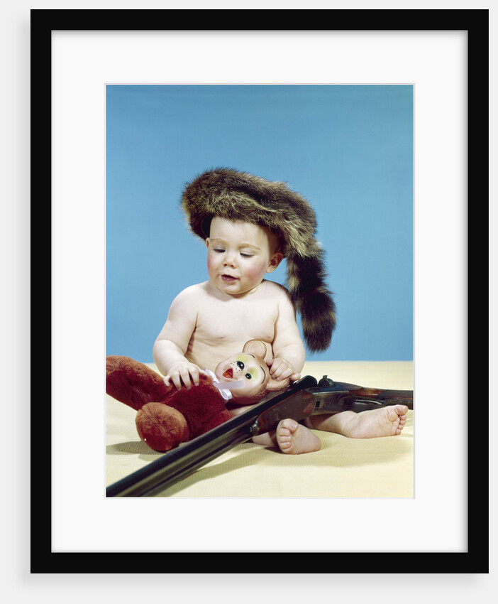 1960s baby boy wearing coonskin cap with stuffed animal and shotgun by Anonymous