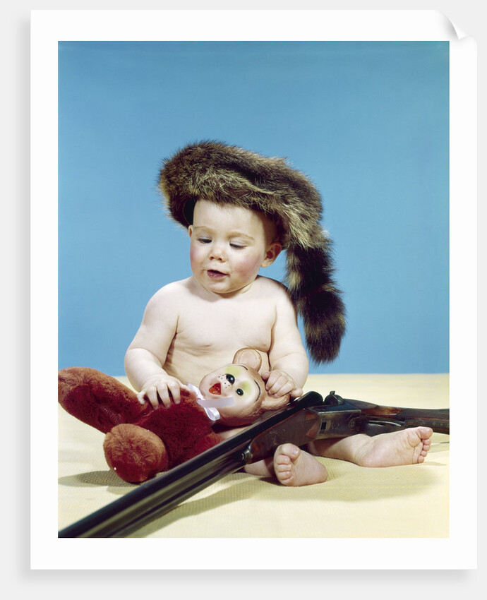 1960s baby boy wearing coonskin cap with stuffed animal and shotgun by Anonymous