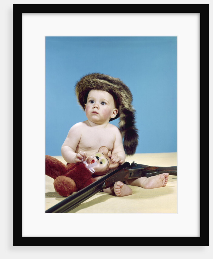 1960s baby boy wearing coonskin cap with stuffed animal and shotgun gun by Anonymous