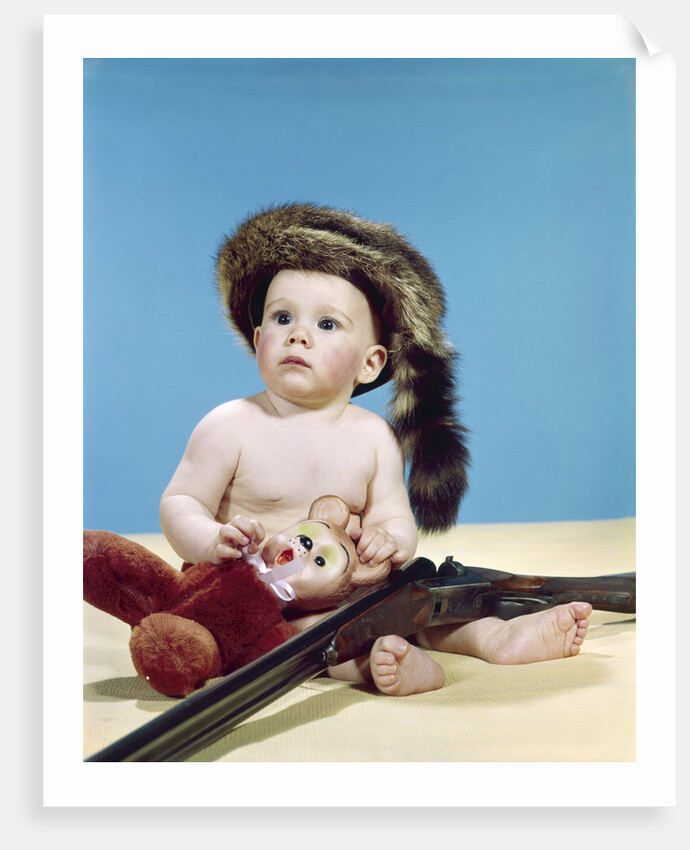 1960s baby boy wearing coonskin cap with stuffed animal and shotgun gun by Anonymous
