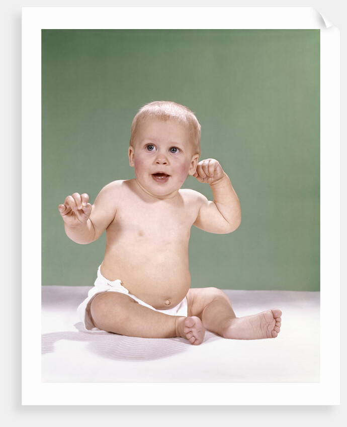 1960s baby wearing diaper sitting on pink blanket mouth open hand touching ear by Anonymous