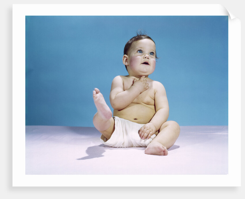 1960s baby wearing diaper sitting looking up and kicking one leg up by Anonymous