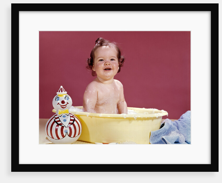 1960s smiling brunette baby sitting in yellow plastic bath tub with toy clown looking at camera by Anonymous