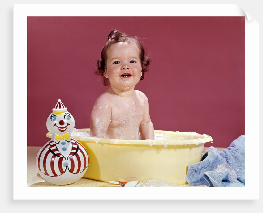 1960s smiling brunette baby sitting in yellow plastic bath tub with toy clown looking at camera by Anonymous