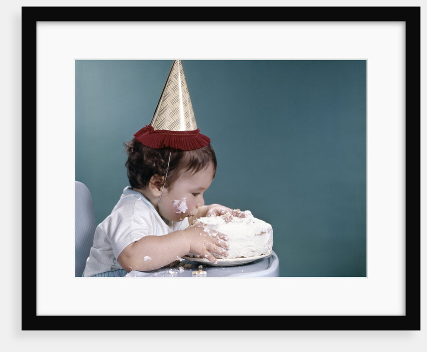 1960s baby in highchair wearing birthday hat eating whole birthday cake by Anonymous