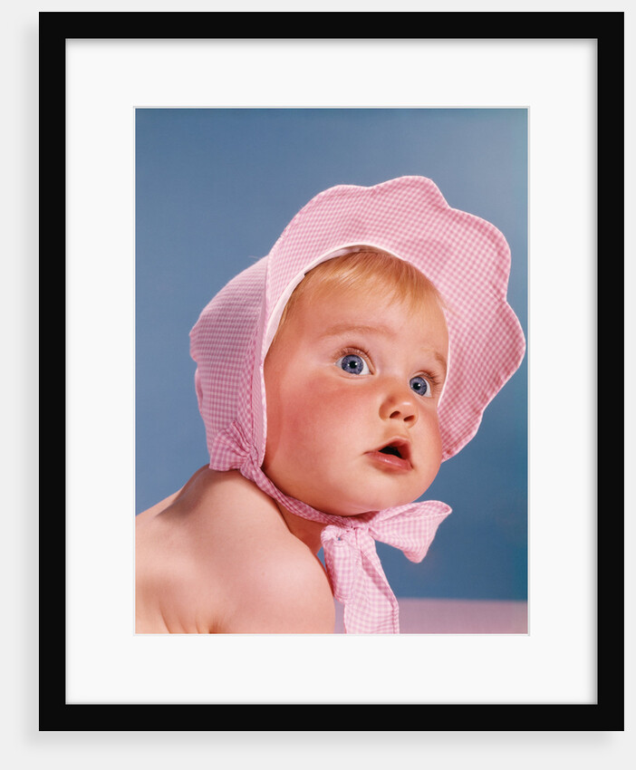 1960s very cute blue eyed baby wearing pink white checked bonnet looking up by Anonymous
