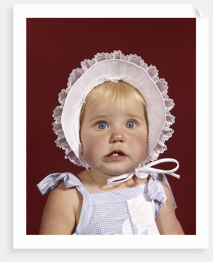 1960s portrait baby girl wearing bonnet and blue gingham dress showing two front teeth looking at camera by Anonymous