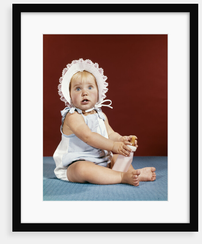 1960s baby girl ruffled bonnet holding bottle funny facial expression looking at camera by Anonymous