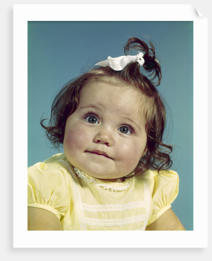 1960s brunette baby girl round chubby face yellow dress ribbon topknot interested sincere facial expression looking at camera by Anonymous