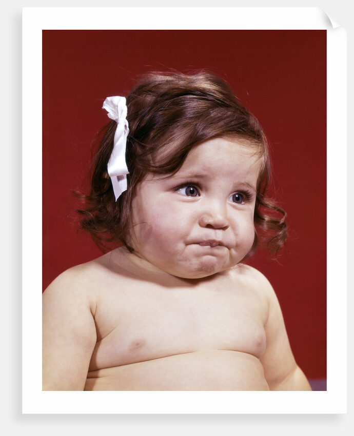 1960s portrait of chubby unhappy baby girl with white ribbon bow in messy hair by Anonymous