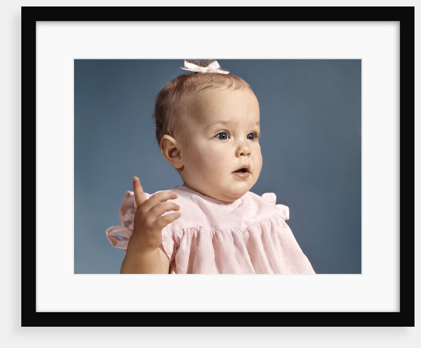 1960s baby girl wearing pink dress with a bow in her hair pointing a finger by Anonymous