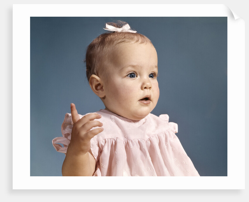 1960s baby girl wearing pink dress with a bow in her hair pointing a finger by Anonymous