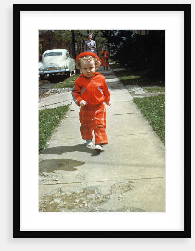 1950s little boy in red outfit running on pavement with mother just behind by Anonymous