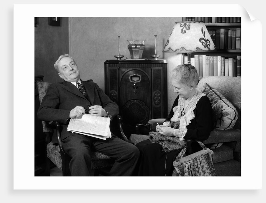 1930s elderly couple sitting in living room listening to radio woman knitting man with newspaper in lap by Anonymous