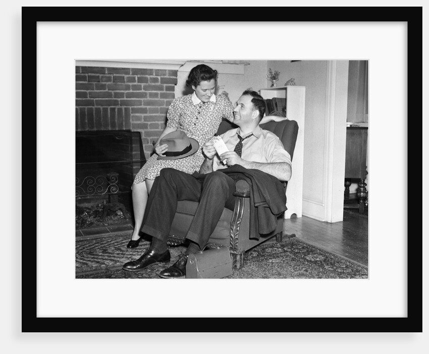1940s smiling man sitting in chair holding pay envelope while wife sits on arm of chair holding his hat lunch box oriental rug by Anonymous