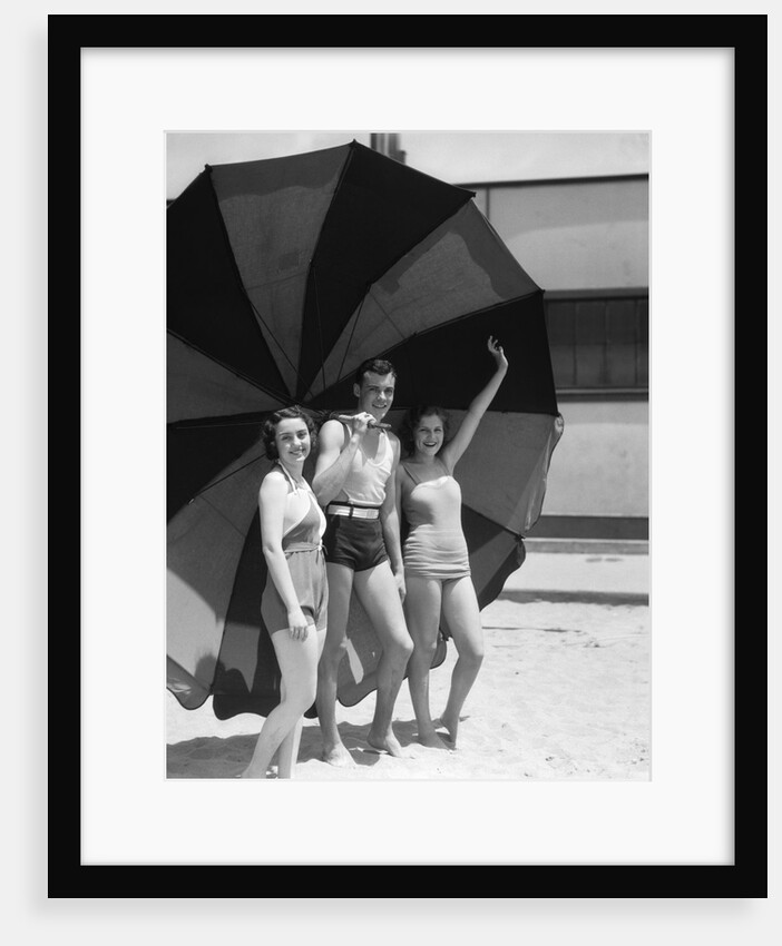 1930s two women one man smiling wearing bathing suits standing under extra large striped beach umbrella by Anonymous