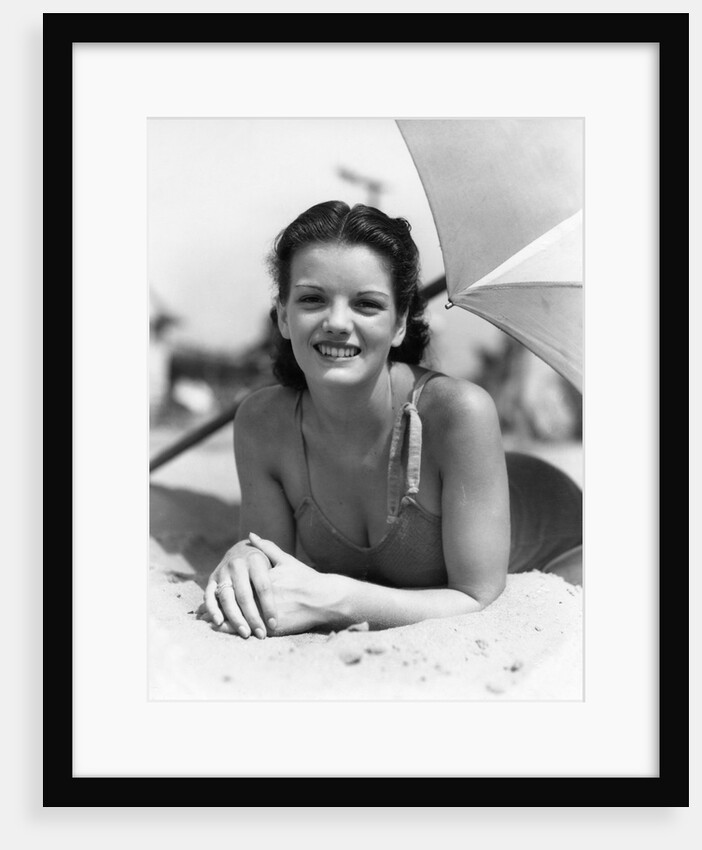 1930s teen girl lying on beach under umbrella wearing bathing suit smiling looking at camera by Anonymous