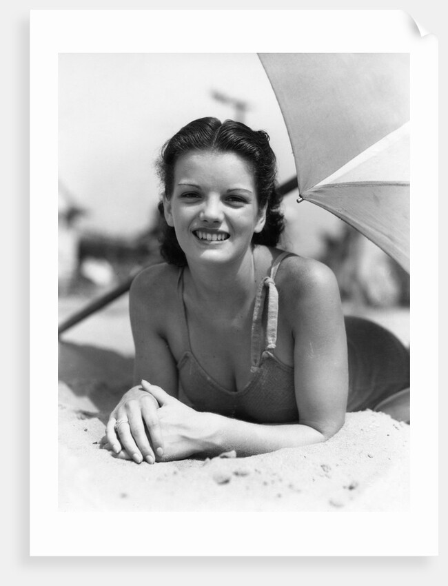1930s teen girl lying on beach under umbrella wearing bathing suit smiling looking at camera by Anonymous