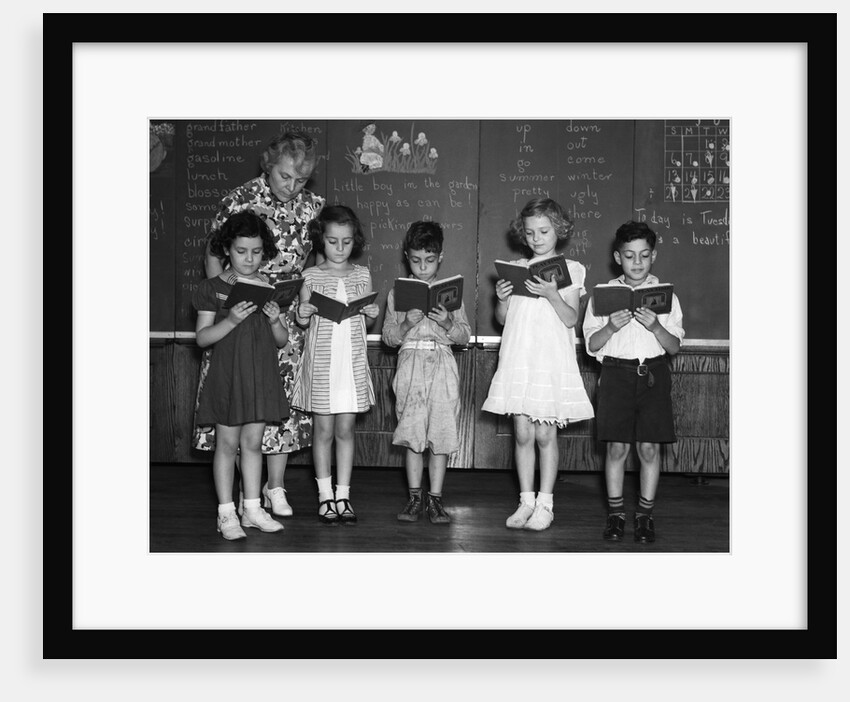 1930s line-up of 5 elementary school students in front of blackboard reading books with teacher looking on by Anonymous